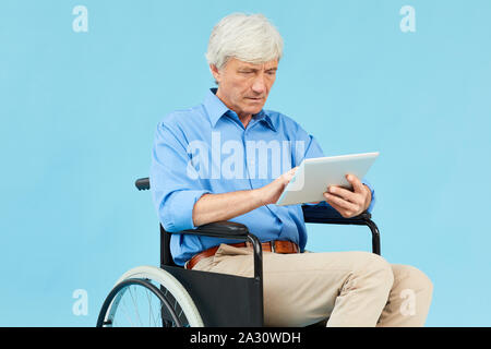 Disabled mature patient sitting in wheelchair and using digital tablet against the blue background Stock Photo