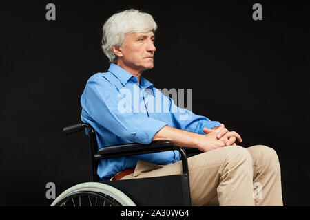 Senior disabled man sitting in wheelchair and looking serious isolated on black background Stock Photo