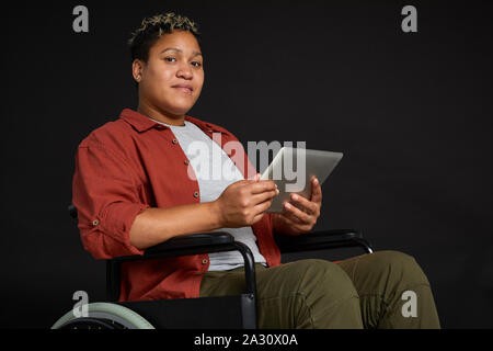 Portrait of African young disabled woman in wheelchair looking at camera while using tablet pc isolated on black background Stock Photo