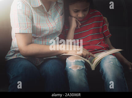Mother and daughter reading book and having fun while spending time together at home under the warm light. Stock Photo