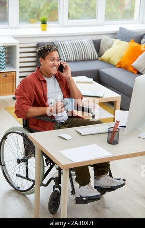Young disabled businesswoman in wheelchair drinking coffee and talking on mobile phone while sitting at her workplace in front of computer Stock Photo
