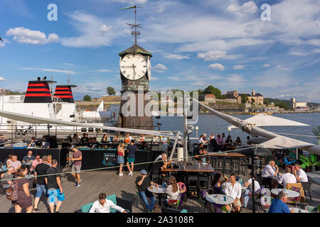 OSLO, NORWAY - People at restaurant on Olso waterfront, and clock tower at Aker Brygge Dock. Stock Photo