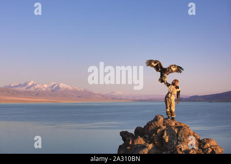 Traditional kazakh eagle hunter with his golden eagle in front of snow capped mountains at a lake shore. Ulgii, Western Mongolia. Stock Photo