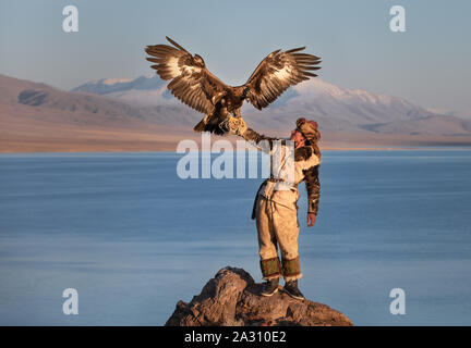 Traditional kazakh eagle hunter with his golden eagle in front of snow capped mountains at a lake shore. Ulgii, Western Mongolia. Stock Photo