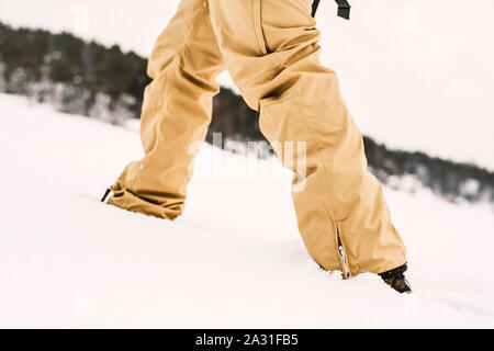 The legs of a man in snowboard trousers, which rises uphill through plump snow, close-up. Stock Photo