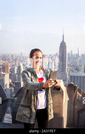 Female tourist takes a selfie on Top of the Rock Observatory with the Empire State Building in the distance, New York City, USA. Stock Photo