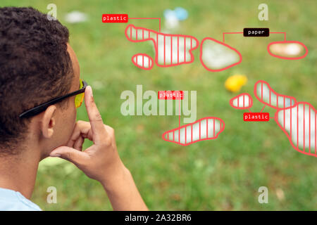 Smart Waste Sorting. African boy wearing digital glasses sitting in the park looking at grass pensive using app for litter management close-up Stock Photo