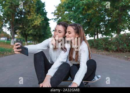 Two girls schoolgirls 13-16 years old, taking pictures phone, summer in city park, background trees fall, sweaters jeans, skateboard. Online call Stock Photo