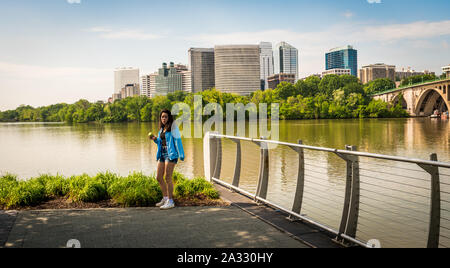 Teen standing by river in blue coat with office buildings in the background Stock Photo