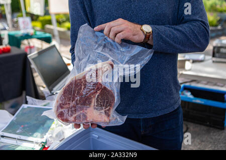 A close up view of a local butcher holding a large t-bone steak, packaged and sold on a market stand during a fair celebrating local farmers.
