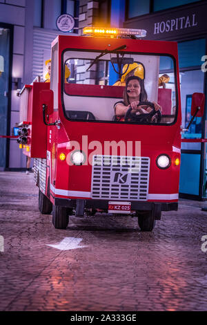 Red firefighter truck with flashing lights driven by a smiling woman is taking a right turn in a mock town of Kidzania Stock Photo