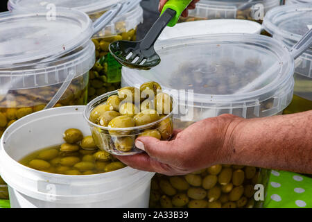 A man's hands are seen up close, filling a small plastic tub with fresh green olives from large pales on a market stand during a local food and produce fair.