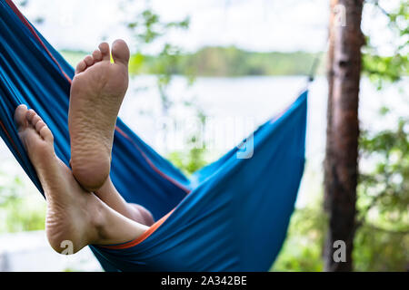 Man's feet are seen close-up from a hammock in a summer forest, on a blurred background of pines and a lake. Stock Photo