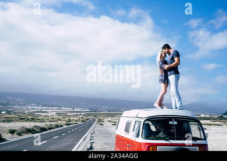 Young couple in love enjoying the travel on a vintage van standing on the roof and hugging with kiss - youth people in outdoor leisure activity and tr Stock Photo