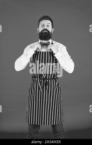 His style works really well to compliment beard. Bearded man fixing bow tie in bib apron. Elegant hipster with bearded face. Bearded bartender or cook in work uniform. Long bearded waiter or servant. Stock Photo