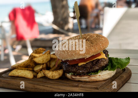 Grilled burger on wooden board with salad and potato chips  with a beach in the background out of focus Stock Photo