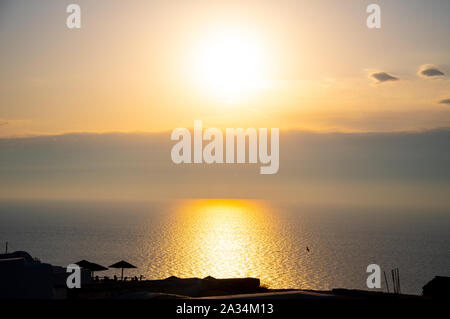 The sun setting behind clouds on the horizon of the Aegean sea seen from a clifftop restaurant in Oia Stock Photo