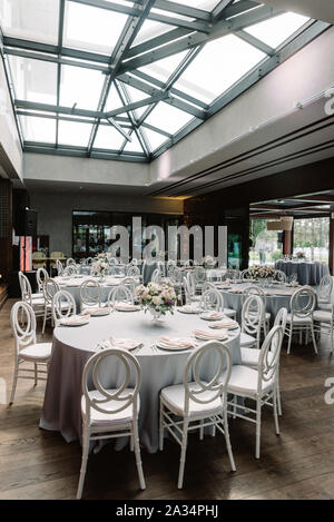 Round holiday tables in the large spacious hall of the restaurant with glass ceilings. Luxurious chairs near round wedding tables. Stock Photo