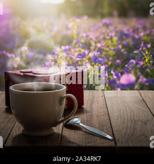 Hot coffee and gift box on wooden table in the meadow of purple flower under sunlight Stock Photo