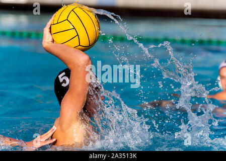 Varisty water polo player in black cap number 5 is bringing up ball for shot on goal with trail of splashing water showing his power. Stock Photo