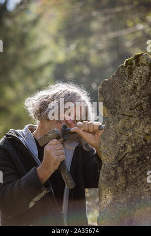 Happy senior sculptor working outside carving in stone using mallet and a chisel. Stock Photo