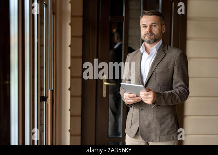 Mature bearded businessman with touchpad looking at you while networking Stock Photo