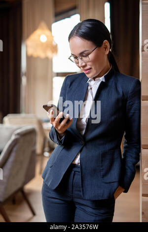 Happy Asian female in elegant suit reading message or scrolling in smartphone Stock Photo