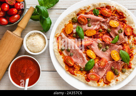 Italian pizza with prosciutto ham, capers, cherry tomatoes, parmesan cheese and decorated with basil leaves. Stock Photo