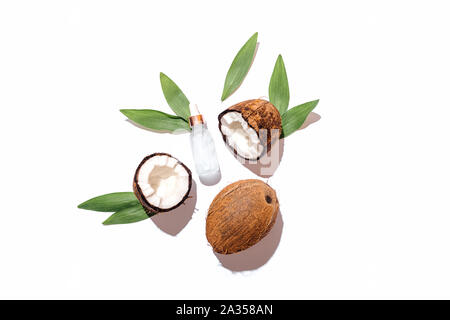 Fresh ripe coconuts with green leaves next to bottle of cosmetic serum on white background, flat lay composition. Stock Photo