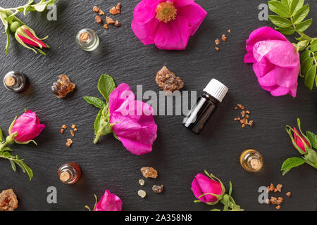 Bottles of essential oil with myrrh, frankincense and Rugosa rose flowers on a dark background, top view Stock Photo