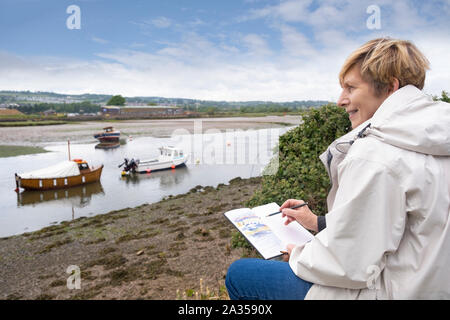 Senior woman enjoying retirement drawing outside near UK river estuary on holiday, wearing casual warm clothing Stock Photo