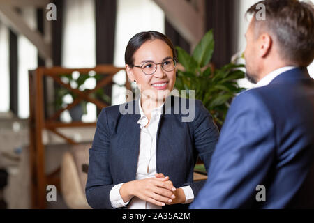 Pretty young cheerful agent in formalwear and eyeglasses talking to client Stock Photo
