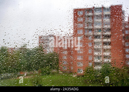 Urban view of rain drops falls on a window during a rainy grey day with green trees and buildings blurred on background Stock Photo