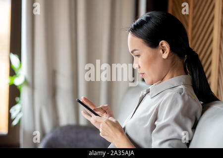 Pretty Asian woman with dark long hair scrolling or texting in smartphone Stock Photo
