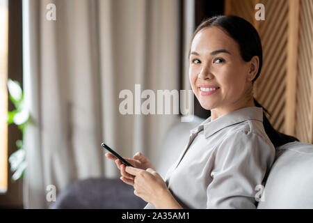 Cheerful young pretty woman in casualwear looking at you while sitting on couch Stock Photo
