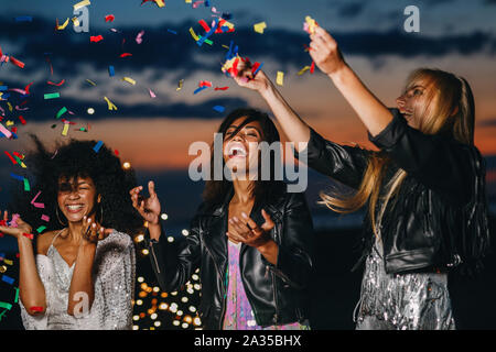 Laughing women throwing confetti at sunset. Three smiling friends having fun together. Stock Photo