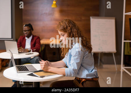 Pretty young casual woman with smartphone texting in college cafe by table Stock Photo