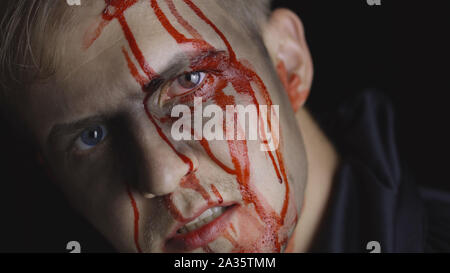 Halloween man portrait with head injury. Guy with dripping blood on his face close-up. Man after fight makeup. Fashion art design. Attractive model in Halloween costume. Dark black background Stock Photo