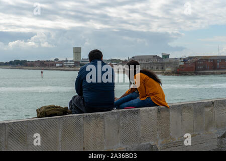 A multicultural couple sitting on a wall at the seaside chatting while looking out to sea at the view Stock Photo