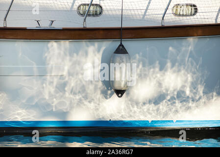 Single white fender hanging on the hull of a sailboat with sparkling reflections of the sun on the water and paintwork Stock Photo
