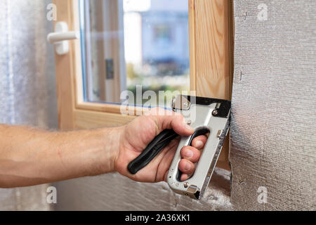A man's hand with the help of a construction stapler fastens onto a wooden beam a foil-insulated heater for the sauna, against the window. Close-up. Stock Photo