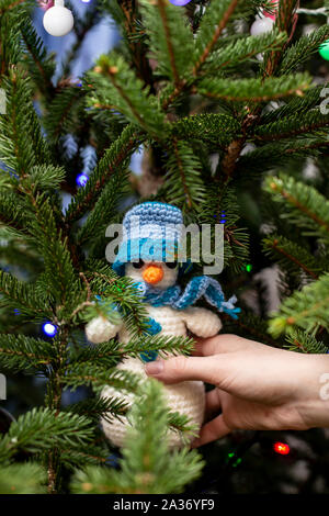 Hand is holding a cute knitted snowman in a hat and scarf on the branches of the Christmas tree, decorated with multi-colored garland. Stock Photo