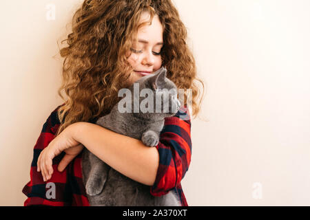 Curly blond teenage girl smiles and holds in her hands a gray kitten Stock Photo