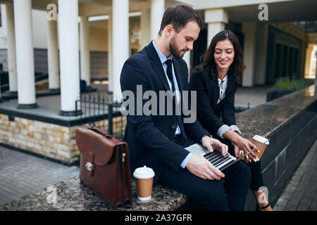 cute handsome man in fashion suit holding laptop ion his knees, reading news from the net while his girlfriend sitting near him and looking at the scr Stock Photo