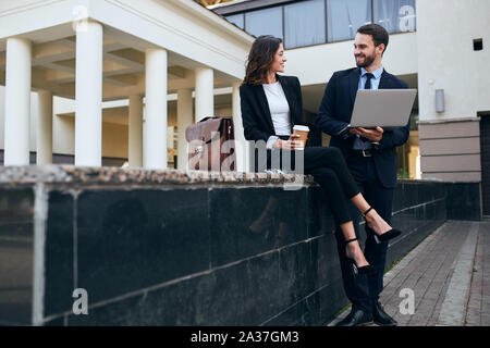 cheerful bearded man holsing a laptop and looking at the woman who is sitting with crossed legs and drinking coffee. copy space Stock Photo