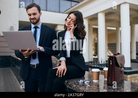 young attractive charming slim woman talking with clients, looking for a sponsors, having a conversation with partners , while male employee using lap Stock Photo