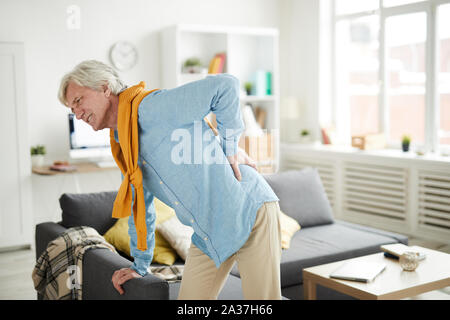 Side view portrait of senior man suffering from back pain and leaning on couch at home, copy space Stock Photo
