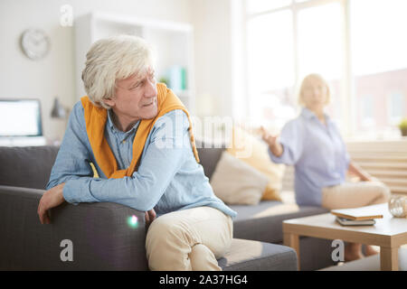 Portrait of senior couple fighting sitting on opposite sides of sofa at home, focus on angry husband in foreground, copy space Stock Photo