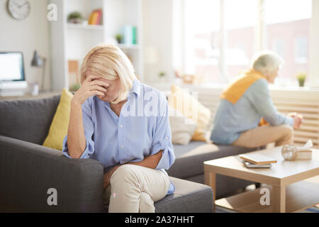 Portrait of senior couple fighting sitting on opposite sides of sofa at home, focus on frustrated wife in foreground, copy space Stock Photo