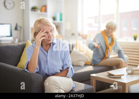 Portrait of senior couple fighting sitting on opposite sides of sofa at home, focus on crying wife in foreground, copy space Stock Photo